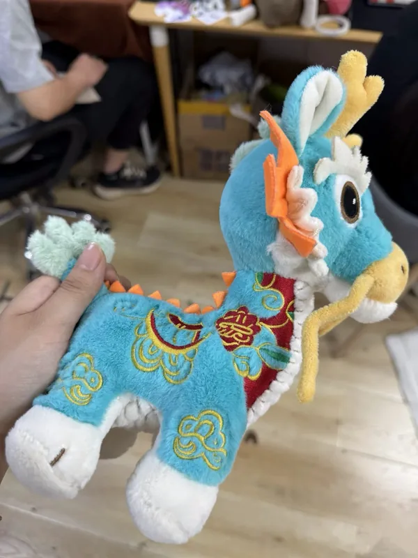 Vue de profil d'un dragon en peluche turquoise avec des broderies dorées et rouges et une longue queue.