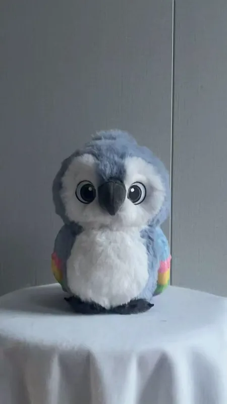 Vue de face d'une peluche en forme d'oiseau gris-bleu, au ventre blanc et aux détails colorés sur les ailes.